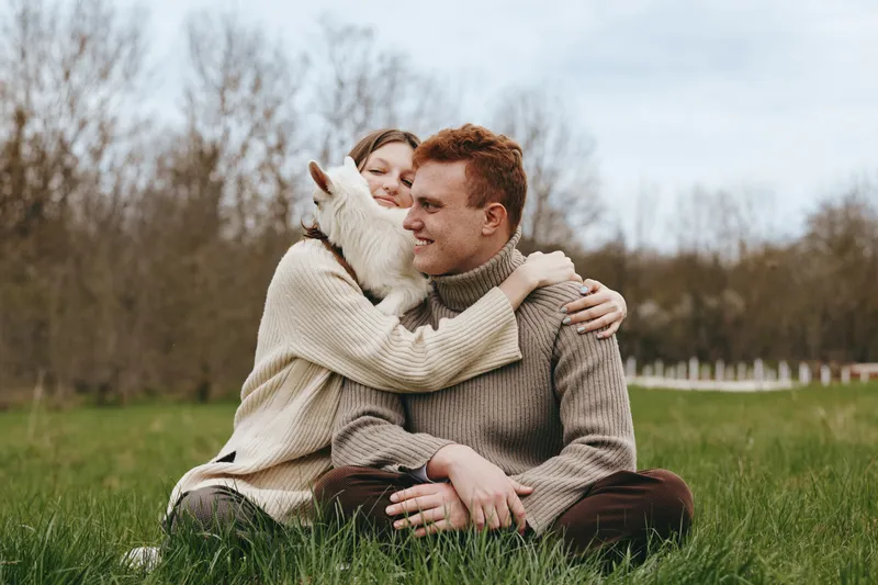 An affectionate couple sits on the grass in a sunny field, sharing a warm hug with a friendly goat kid between them. A candid lifestyle moment capturing connection, nature, and rural life.