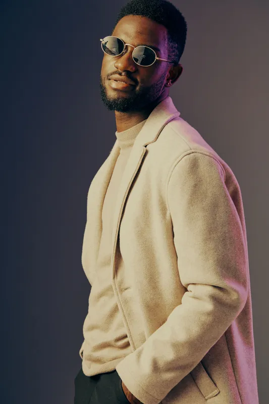 Stylish young Black man in sunglasses wearing a beige coat against a gradient background, showcasing modern fashion and confidence