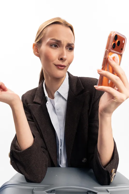 Young woman in blazer looks confused and doubtful while holding smartphone. Casual indoor setting with white background. Concept of technology frustration and communication issues.