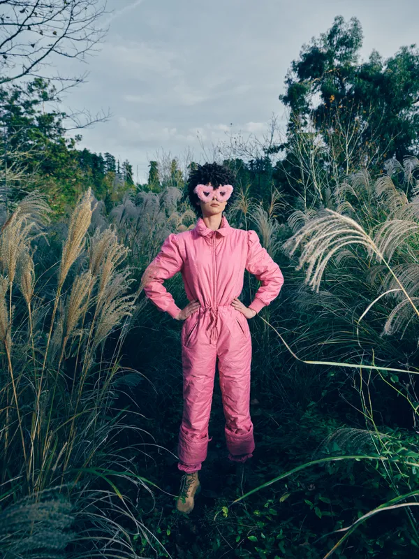 Person, pink jumpsuit, mask, outdoors, tall grasses, garden setting, fashion, bold stance, creative character, vivid color, whimsical mood