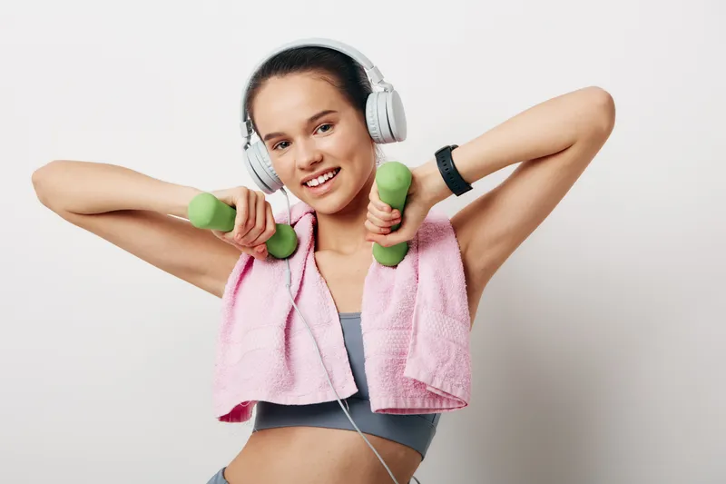 Young woman fitness portrait showing a smiling female in activewear with pink towel over shoulders, lifting green dumbbells while wearing headphones against a clean white studio background.