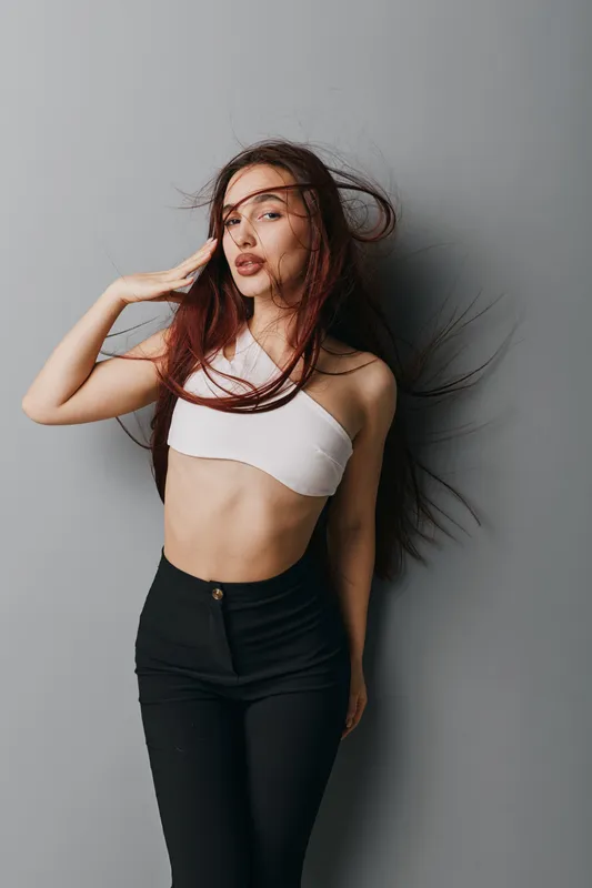 Young woman with long hair posing against gray background wearing white crop top and black pants. Fashion model with natural makeup and casual style in studio for portrait photography.
