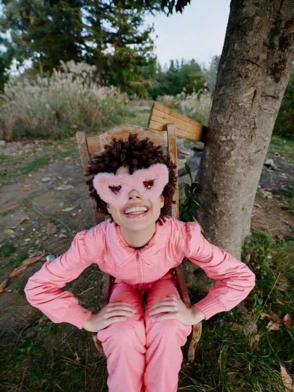 Child girl in pink costume wearing a fluffy heart-eyed mask, sitting on a wooden chair outdoors, smiling with hands on hips in a sunny nature garden scene