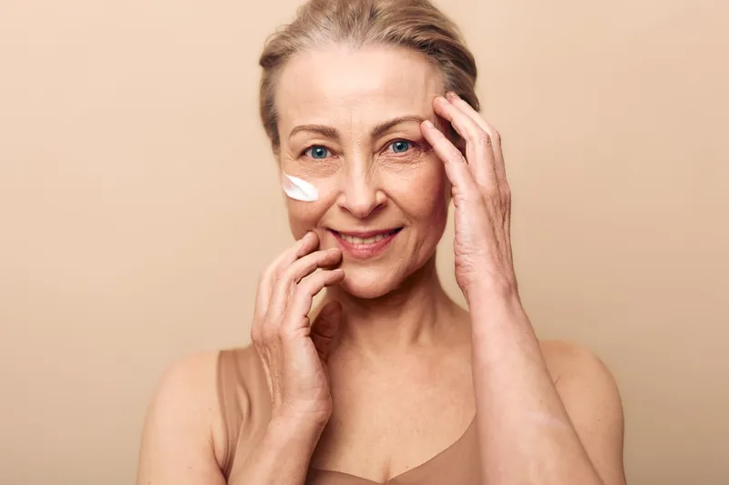 Mature woman with blue eyes applies white face cream on cheek, smiling gently. Close-up on beige background. Concept of skincare, aging skin care, self-care, and natural beauty routines.