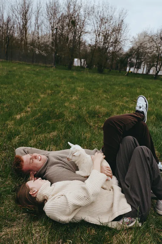 Casual couple lying on grass embraces a small goat kid in a sunny park, capturing a warm lifestyle moment of affection, relaxation, and animal companionship amid soft sweaters and simple nature.