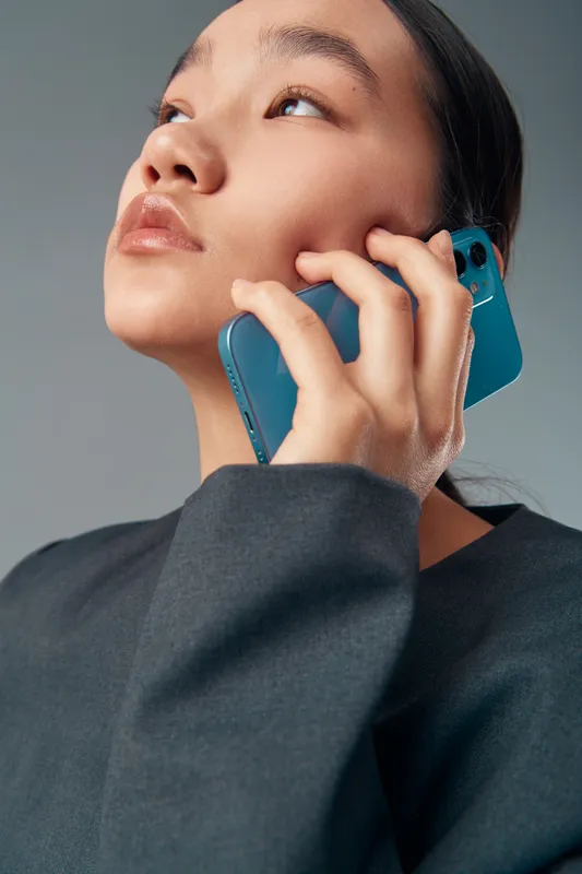 Young woman holding blue smartphone close to face speaking with focus. Portrait, communication, technology and person wearing dark clothing on casual gray background.