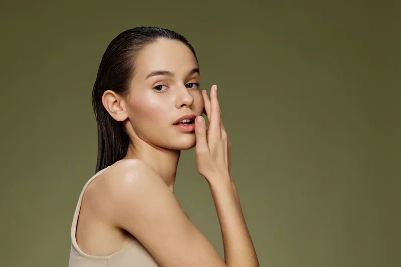 Young woman with flawless skin touching her face, showcasing a natural beauty concept against a soft green background, ideal for skincare and beauty promotions