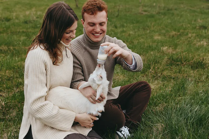 Couple outdoors in a meadow with a furry goat kid, smiling as the man feeds milk, the woman comforting, a tender love filled lifestyle moment in nature, rustic pasture scene with grass and calm