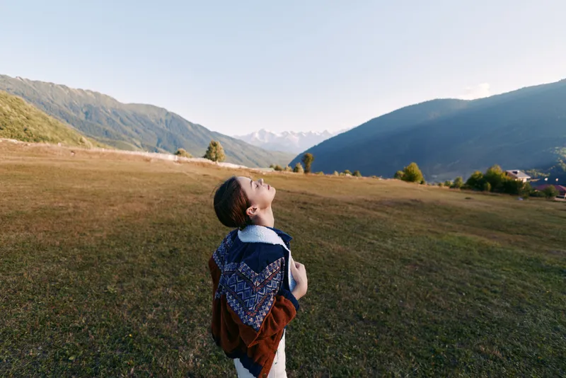 Girl with backpack standing in sunlit meadow looking up at distant mountains and valley, enjoying nature and rural landscape, freedom, exploration and a peaceful outdoor moment.