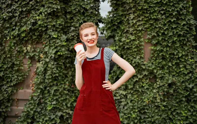 Симпатичная женщина с чашкой кофе рядом с зелёными листьями кустов на улице