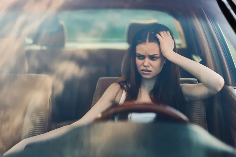 Young woman showing frustration while driving in a car, with a worried expression, blurred background, and natural lighting capturing a seasonal moment.