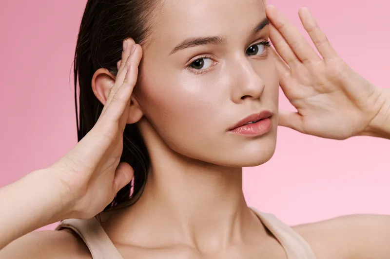 Young woman with natural beauty showcasing healthy skin, neutral attire, and subtle emotions, against a soft pink background, emphasizing skincare and wellness concepts