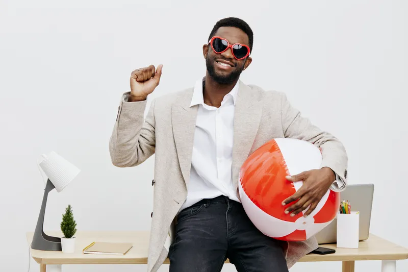 Joyful young man with dark skin, wearing red sunglasses and a beige blazer, holding a colorful beach ball and smiling broadly, standing in a modern office with white walls and minimal decor. People li