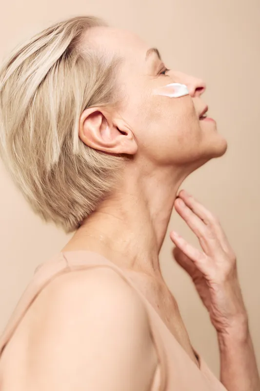 Profile of mature woman with short blonde hair applying face cream, showing smooth skin and gentle smile. Concept of skincare, aging beauty, self-care, and natural wellness.