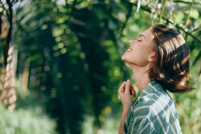 Woman enjoys the sunshine as she tilts her head back, eyes closed in contentment, surrounded by lush greenery and dappled light, capturing peaceful outdoor serenity and gentle warmth.