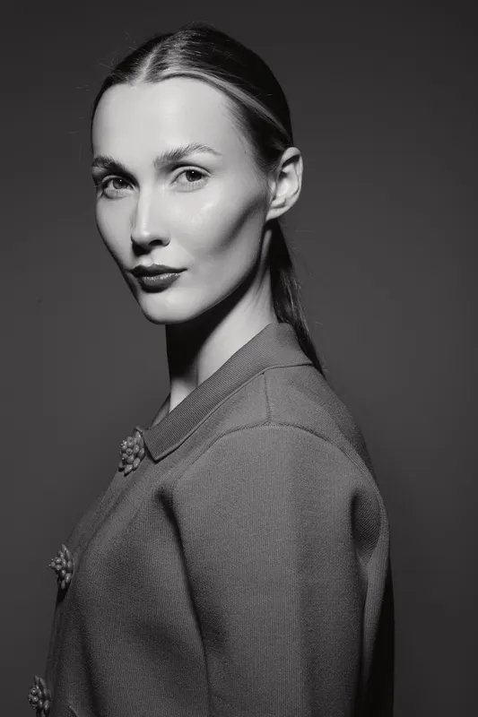 Portrait of a young Caucasian woman with fair skin and slicked back hair wearing a textured jacket in a black and white studio setting.