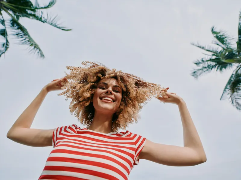 Joyful young woman with curly hair in a red and white striped shirt smiles toward the sky, arms lifted under bright daylight beside palm trees in a sunny outdoor setting, exuding energy and carefree