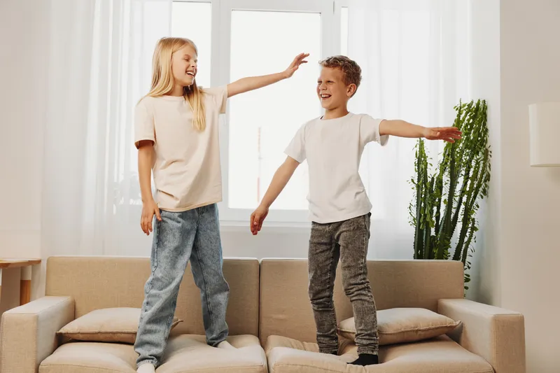 Two happy children standing on a beige sofa in a bright living room, smiling and playing together. Casual clothes and natural light create a warm, joyful home atmosphere.