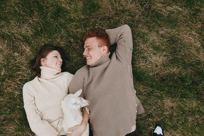 Cheerful couple outdoors, lying on grass with a playful goat kid nearby, wearing cozy sweaters, warm sunlight, relaxed embrace, soft breeze, candid lifestyle moment, natural colors