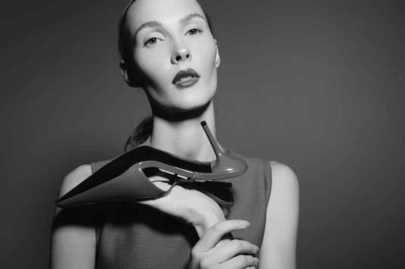 A pale Caucasian woman with slicked-back hair holds a high heel shoe near her face, posing with a serious expression in dramatic black and white studio lighting. Concept of fashion, elegance, and