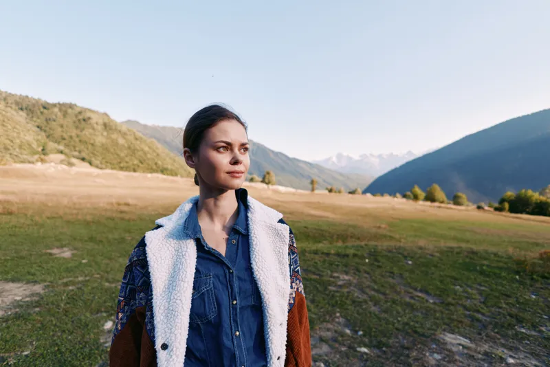 Woman portrait in mountains meadow, nature outdoors landscape scene with casual denim shirt and shearling jacket, thoughtful traveler enjoying open field and distant peaks and soft golden light at