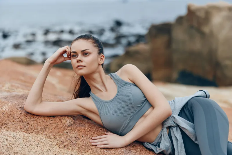 Young fitness model in sporty attire relaxing on rocky beach, showcasing healthy lifestyle, serene expression, and natural beauty