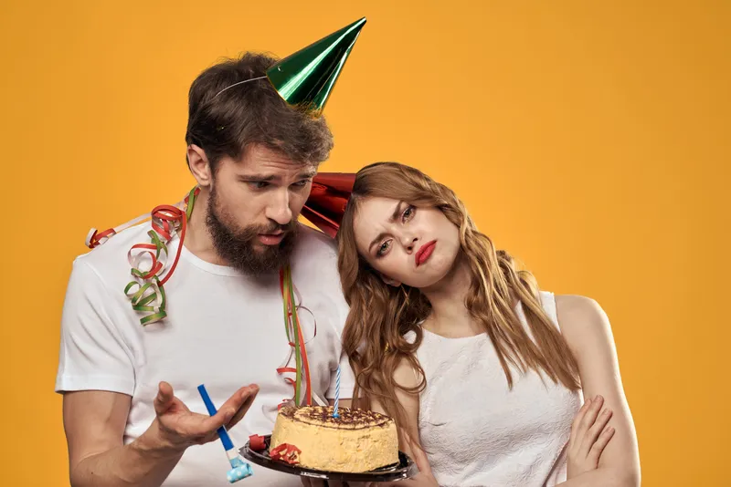 Мужчина и женщина, праздничный торт на день рождения, жёлтый фон и колпаки на головах