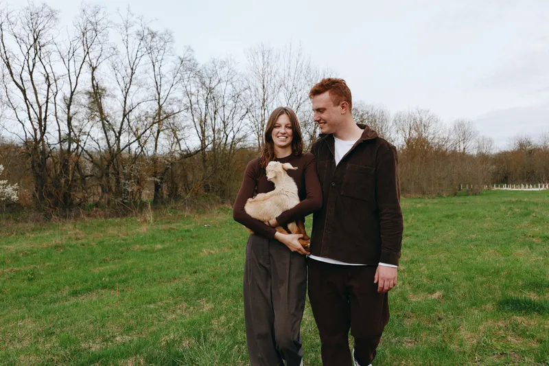 Smiling couple walking through a grassy field, holding a fluffy goat kid. Natural light and open countryside convey warmth, connection, and a simple rural lifestyle.