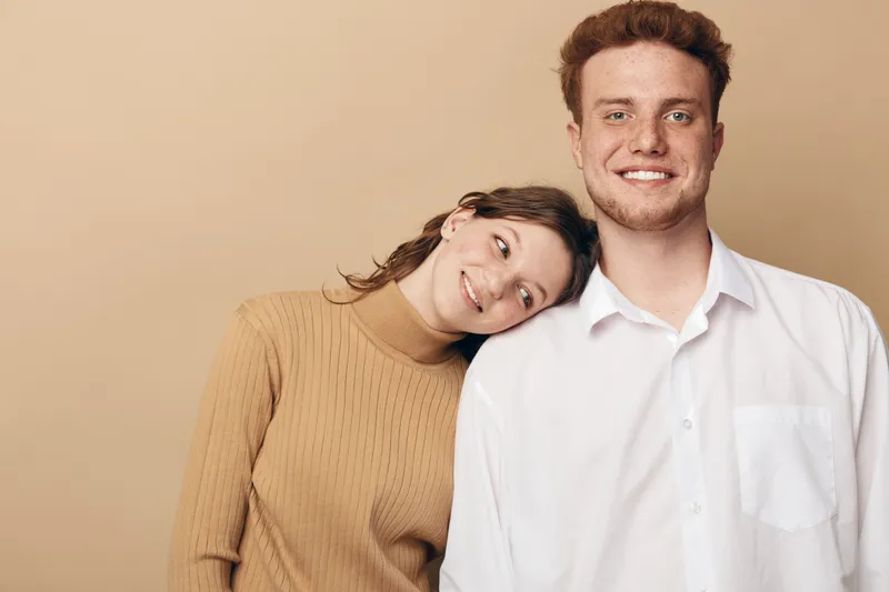 Smiling young woman in beige sweater leans on shoulder of happy man in white shirt. Warm studio lighting and neutral background create cozy, intimate atmosphere of friendship and affection.