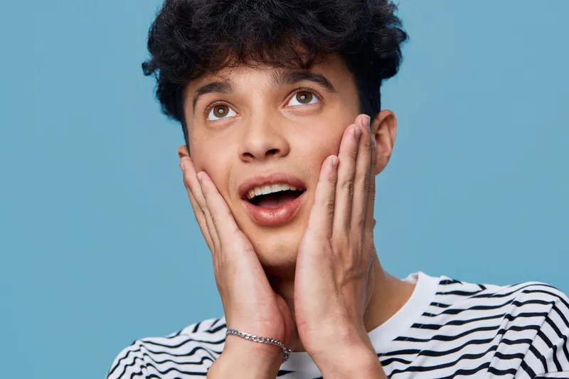 young man with surprised expression touches his face looking upwards against blue background, casual striped shirt and curly hair convey curiosity and astonishment