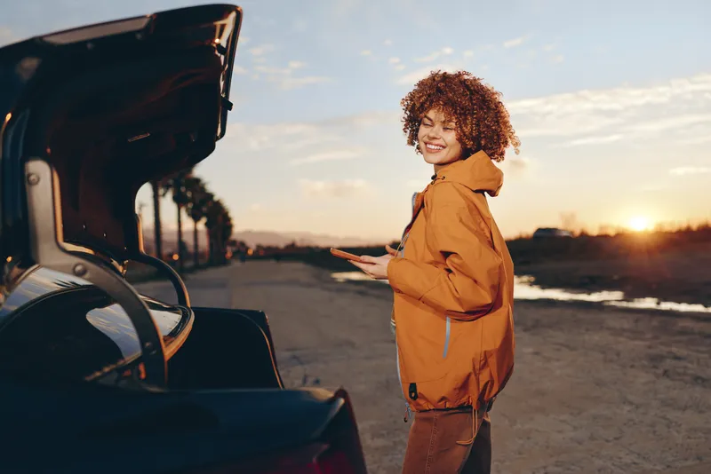 Woman lifestyle outdoors at sunset smiling curly hair evening portrait near open car trunk orange jacket warm golden sunlight soft breeze relaxed stance