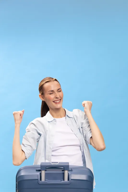 Happy young woman with suitcase celebrating success on blue background. Travel concept with casual outfit, joyful expression, and positive mood for vacation or journey planning.