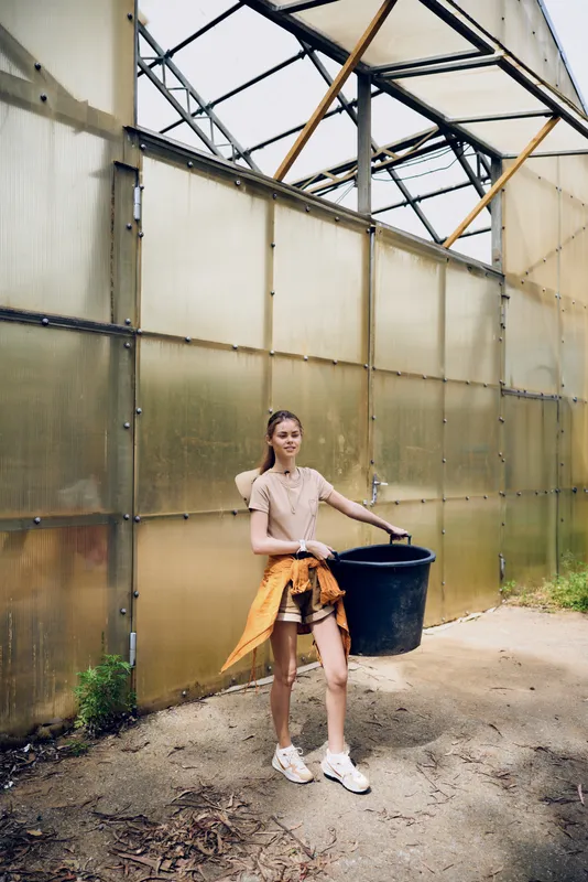 Girl teenager in greenhouse carrying a black bucket for gardening, casual summer outfit and sneakers, plants and soil work in a bright nursery with structural glass panels and sunlight.