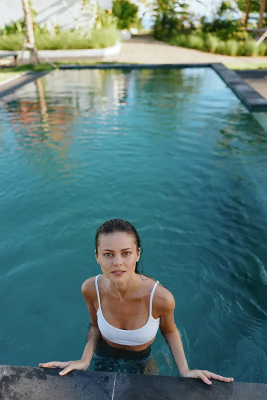 Smiling woman in a white bikini stands at the pool edge, hands gripping the wall. Outdoor scene highlights lifestyle, fitness, and summer refreshment with warm sunlight and calm water.