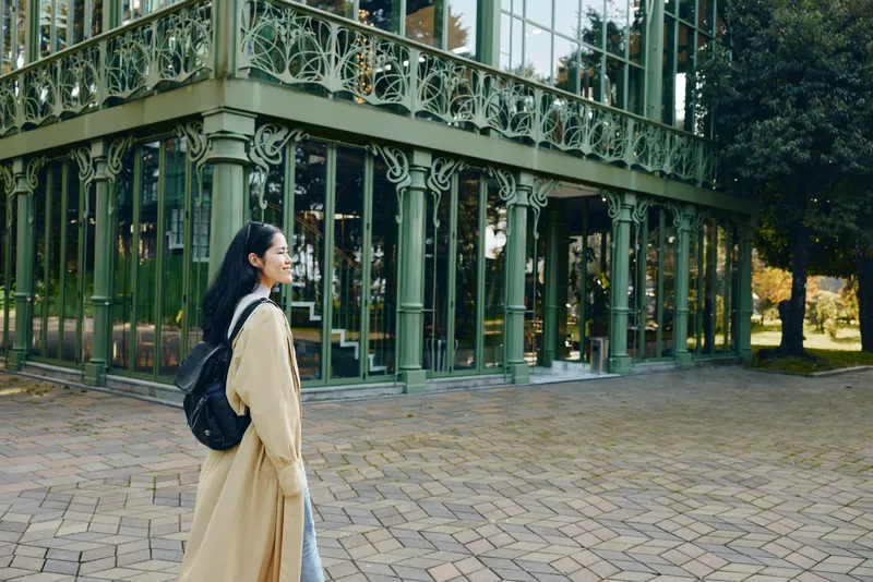 Woman in beige trench coat with backpack walking near glass architecture in an urban courtyard, daytime stroll, calm atmosphere, fashion forward street scene