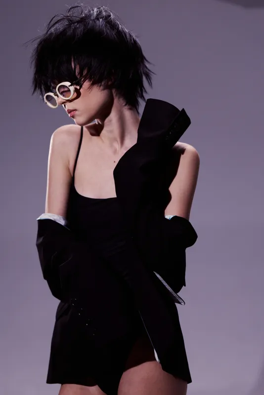 Stylish woman with short black hair wearing sunglasses and black outfit in moody studio lighting.