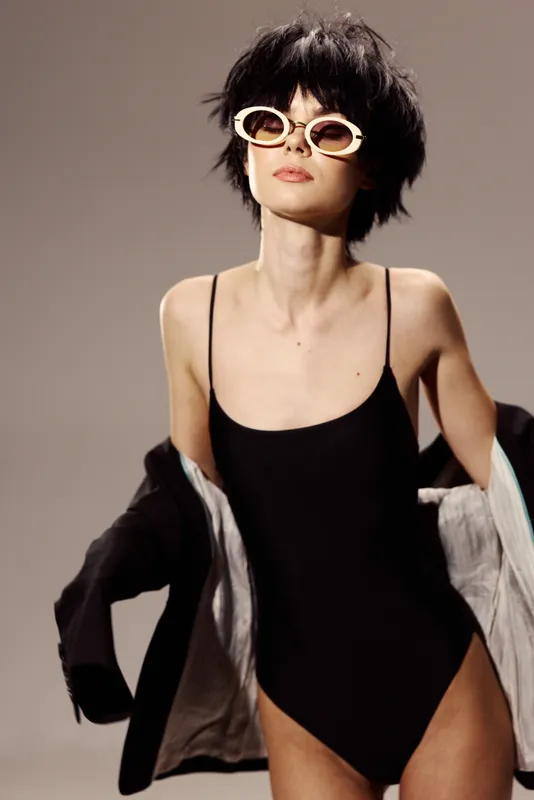 Stylish woman in black swimsuit and sunglasses posing with a jacket on beige background, fashion, confident, summer, trendy look.