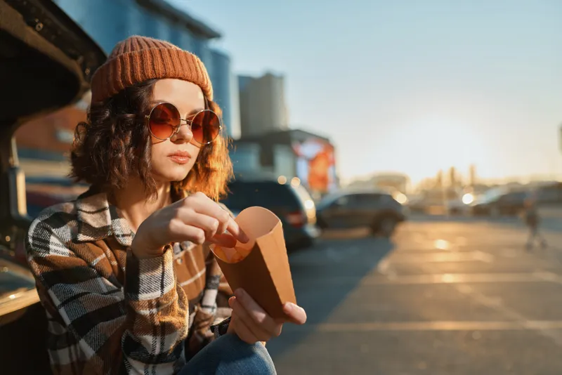 Woman eating fries from a car trunk in a parking lot at sunset, wearing beanie and sunglasses. Candid lifestyle portrait with authenticity, golden hour glow, mindful living and emotional storytelling.