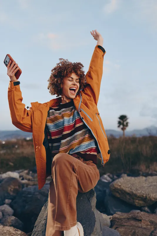 Woman lifestyle outdoors, smiling with curly hair, wearing an orange jacket, holding a smartphone, arms raised on a rocky trail, backpack visible, adventure under open sky, morning light