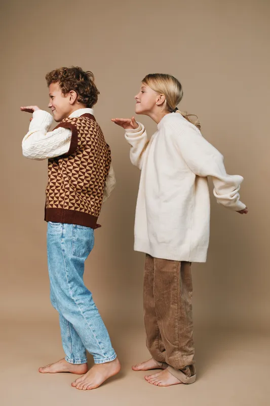 Two barefoot adults pose playfully, side by side, one in a patterned knit vest and jeans, the other in a cozy oversized sweater and soft pants, miming a whistle.