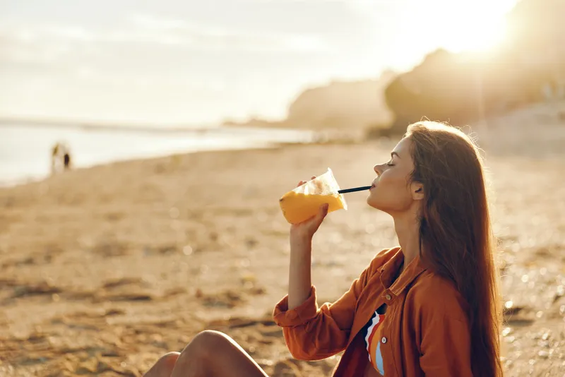 Woman, beach, sunset, drinking, juice, carton, outdoors, sunlight, relaxation, sitting, profile view, young woman smiles, hydrating from carton, warm summer breeze, calm sea waves, sandy shore