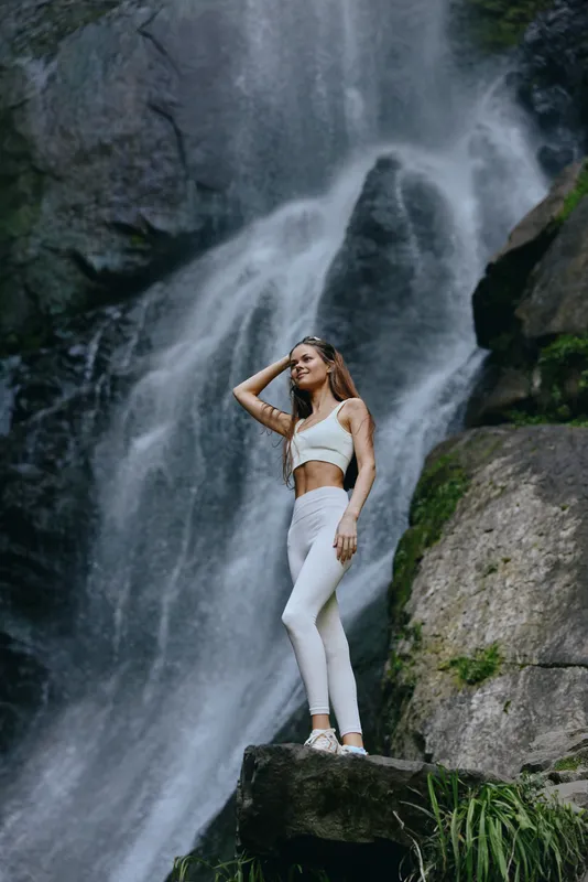 woman standing near waterfall in white athletic wear looking up thoughtfully, natural outdoor scene with rocks and flowing water, fitness and adventure mood