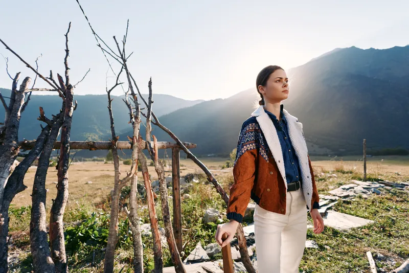 Woman in jacket standing in a rural field with mountains behind, sunlight on her face, looking thoughtful. Scenic landscape, casual outfit, wooden fence and open sky at golden hour