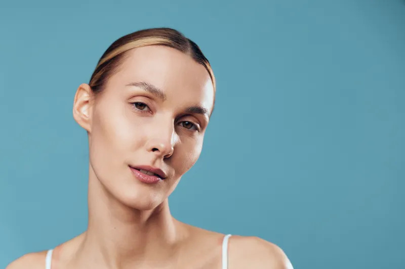 Young woman with clear skin and natural makeup posing against blue background, soft lighting, minimalistic style, calm expression, smooth complexion, close-up portrait