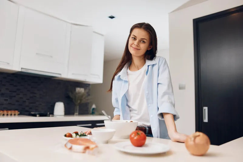 Молодая женщина стоит перед кухонной стойкой с миской свежих фруктов и овощей
