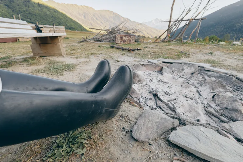 Boots resting beside a cold campfire with grey ash on the ground, mountains in the background and outdoor countryside campsite scene with bench, wooden firepit and rugged landscape.