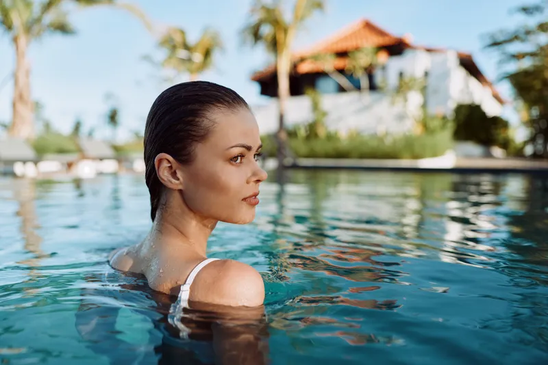 Woman in a pool, side profile, tropical backdrop, relaxed mood. Clear water, warm sun, and calm reflections convey leisure, wellness, and lifestyle in a vacation setting.