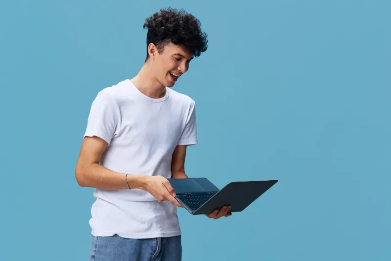 young man holding laptop smiling in casual white t-shirt and jeans with blue background technology and communication concept