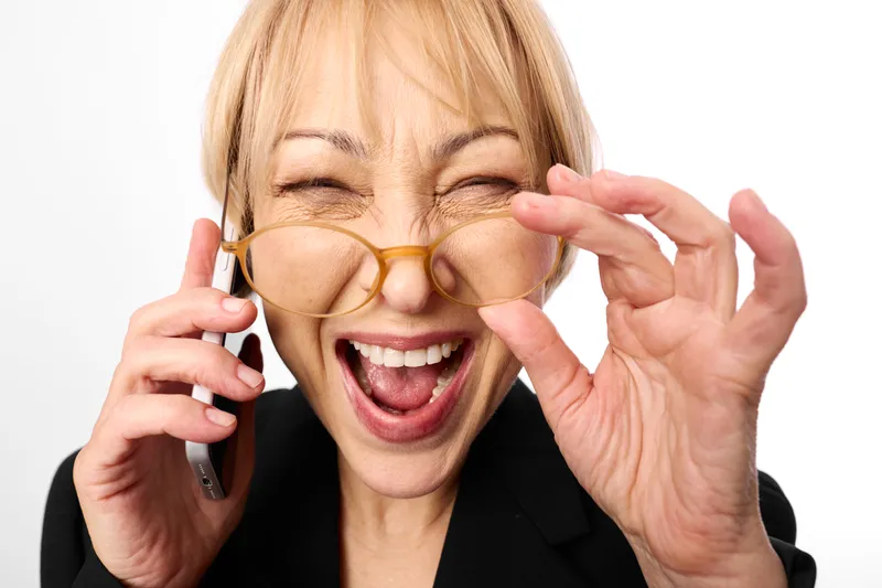 Woman glasses smile phone blonde laughing adjusting eyewear with hands on white studio background, bright natural light, playful expression, minimal scene, business call moment