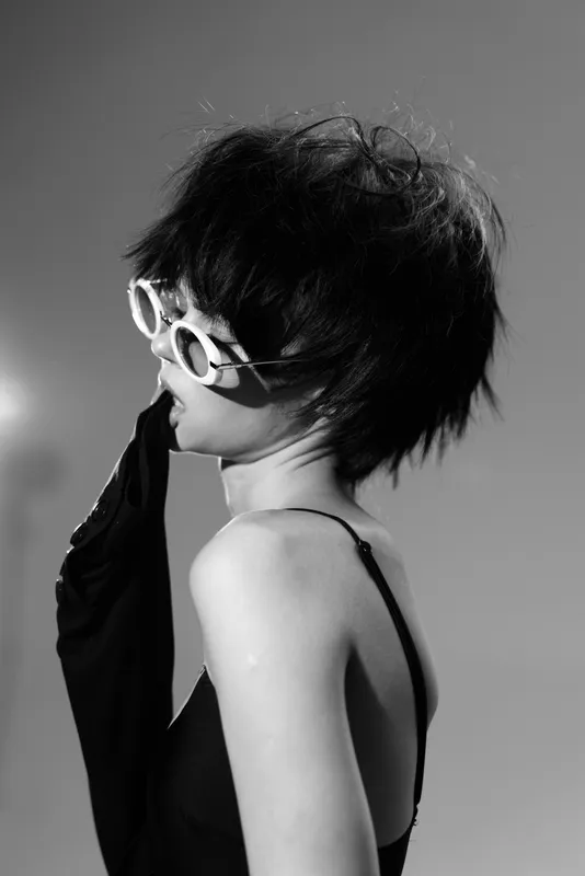 Black and white portrait of a stylish woman wearing sunglasses and gloves with dramatic lighting and moody atmosphere.